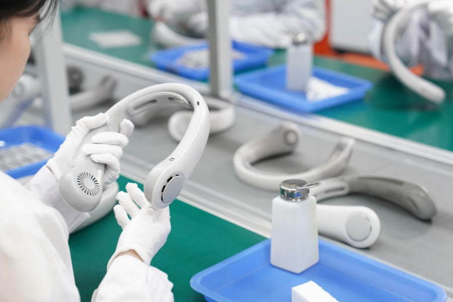 A quality control worker in a cleanroom environment carefully inspects a newly assembled white plastic neck fan on a green manufacturing assembly line.