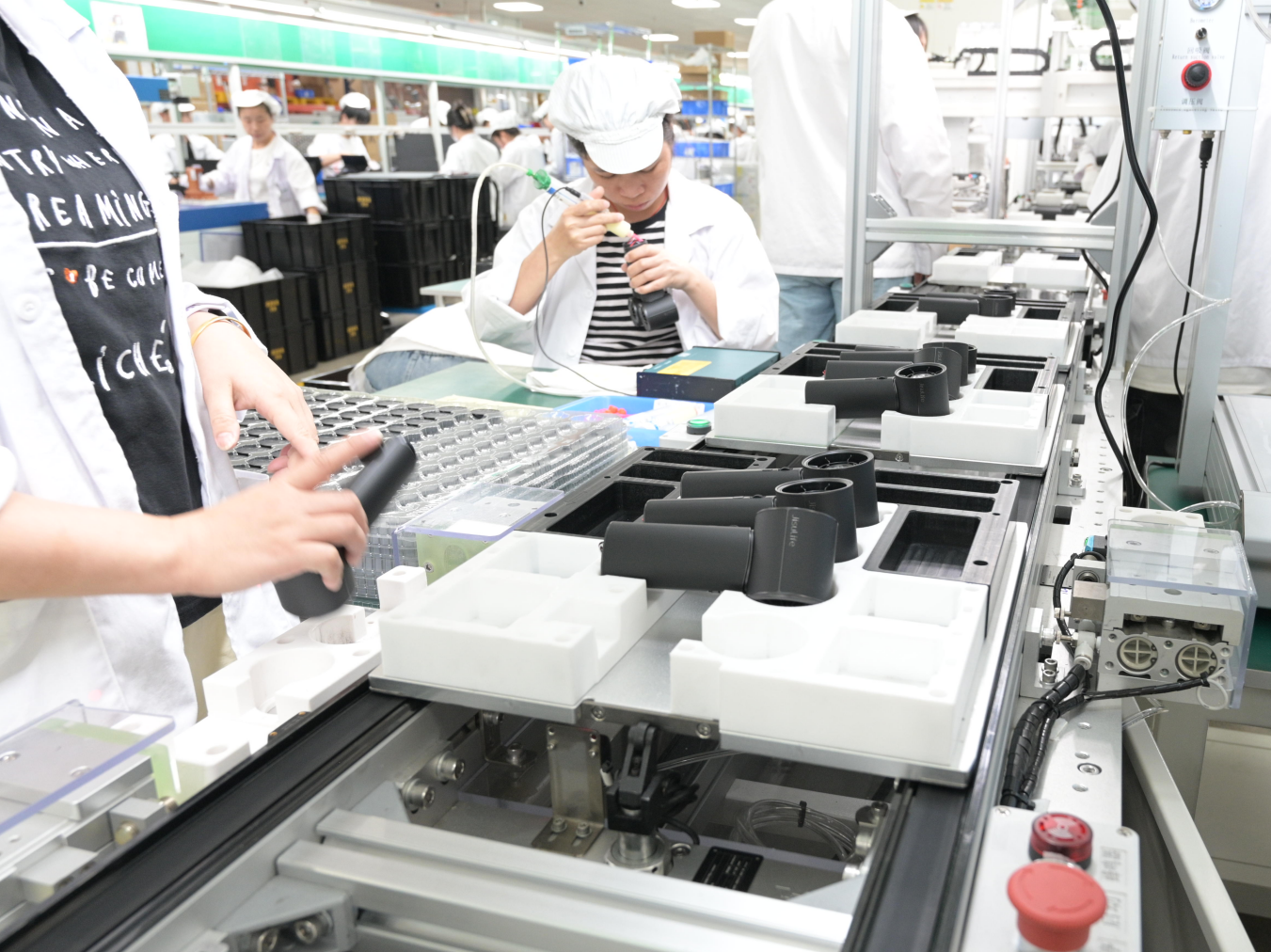 Workers assemble black plastic components on a production line in a manufacturing facility.
