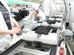 Workers assemble black plastic components on a production line in a manufacturing facility.