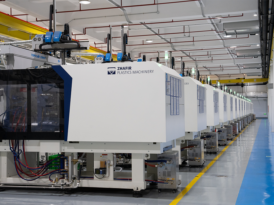Rows of clean white injection molding machines line a bright factory floor with overhead cranes and organized production equipment nearby.