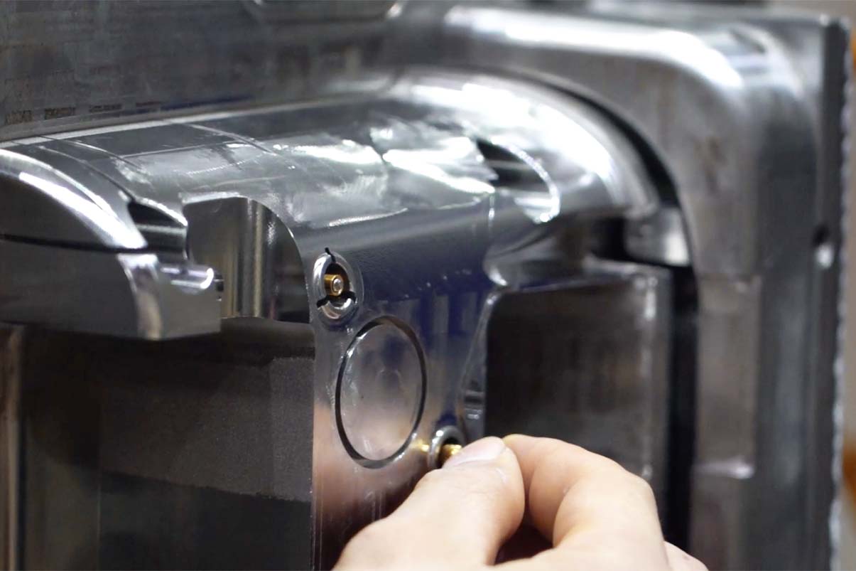 A close-up view of a technician's hand carefully placing a small brass insert into a highly polished metal injection mold cavity.
